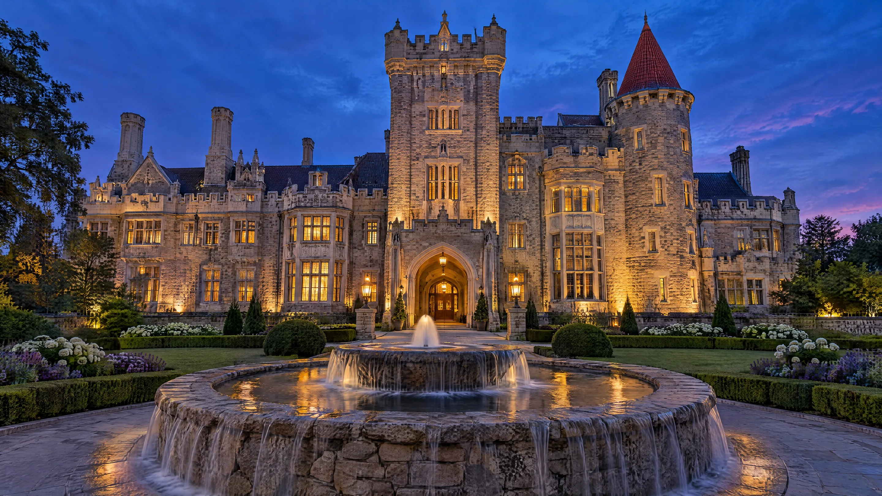 An illustrated Gothic Revival castle lit at dusk behind a stone fountain and gardens